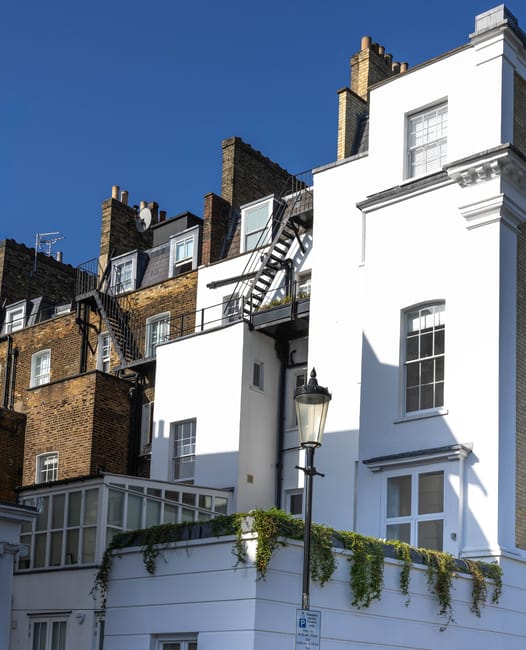 A group of brown brick and white-painted residential buildings in an urban setting, with external fire escape staircases attached to the facades, some leading to rooftop access. The buildings feature multiple windows, including dormer windows on the upper floors, and rooftop satellite dishes. The bottom part of the image shows a white ground-floor extension with large windows and decorative greenery hanging over the edge. A black street lamp with a glass lantern is positioned in front of the buildings, casting a shadow on the white facade, all under a clear blue sky. This scene depicts typical city housing and balcony access, relevant to house removals, relocation services, and staircases access, aligning with advice on access issues for Highbury flats and staircases removals, as seen on highburymanandvan.co.uk.