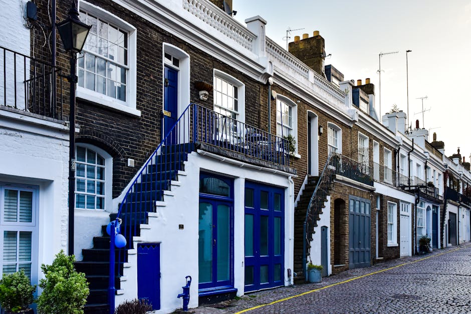 Exterior view of a row of terraced houses with brick facades and white decorative trim, taken during daylight with overcast sky. The image shows the ground floors and upper balconies of the properties. In the foreground, a house has a prominent blue front door with a large glass window, flanked by a blue-framed window on the left. A metal staircase painted in matching blue ascends from the sidewalk to a small balcony with metal railings in front of the upper door, with a potted plant visible on the balcony. The buildings are positioned along a cobbled street with a yellow line marking the edge of the pavement. Potted plants and small shrubs are placed near the entrances. A black outdoor lamp is mounted on the wall next to the staircase, and various chimney stacks and antennas are seen atop the roofs. The scene captures elements relevant to house removals and furniture transportation, highlighting the importance of accessible entry points for home relocation services provided by Highbury Man and Van.