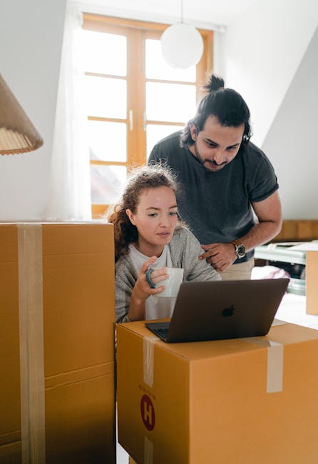 A man and a young girl are inside a home, engaged in packing and moving preparations. The girl, seated at a wooden table or surface, is holding a white mug while looking at a laptop computer placed on top of a large cardboard box, which is sealed with clear packing tape. The man, standing beside her, leans slightly forward, observing the screen, and has dark hair tied back in a bun, wearing a dark t-shirt. Around them, there are several cardboard boxes, some stacked behind and beside them, suggesting an active house relocation process. The room is well-lit with natural sunlight streaming through a large window with wooden frames and a white ceiling light hanging overhead. This scene reflects the early stages of home relocation, involving packing items, digital planning, and coordinating household moves, as handled by Highbury Man and Van, specialists in removals and transport services.