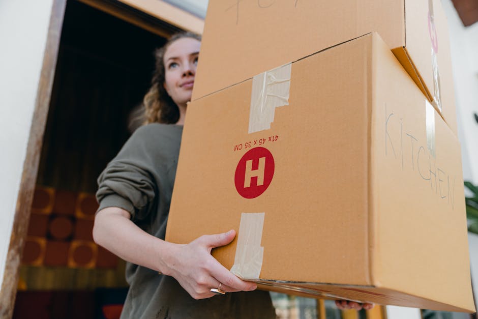 A woman with long, wavy brown hair holding a large cardboard box labeled with a red 'H' in a house interior, with the woman positioned near a doorway. The box appears to contain household items, possibly for moving, and is sealed with packing tape. Scribbles and writing are visible on the box, indicating contents or destination. In the background, there are stacked items and part of a wooden doorframe, suggesting a home environment during a furniture transport or home relocation process. The lighting appears natural, highlighting the careful handling of the packed box, which is part of a moving or packing and moving service offered by Highbury Man and Van, a specialist in removals and home relocation.
