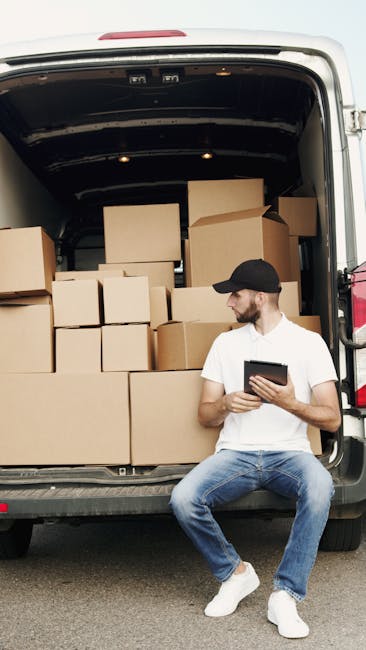 A young man with a beard, wearing a white t-shirt, blue jeans, white sneakers, and a black cap, is sitting on the pavement next to an open commercial van involved in a home relocation process. Inside the van, there are numerous brown cardboard boxes of various sizes, some stacked on top of each other, and others placed on the vehicle's floor. The boxes are sealed with packing tape, indicating they have been packed for moving. The interior of the van is dark and spacious, with a black ceiling and walls. The man is holding a tablet, possibly coordinating or checking the loading process, while the boxes are being prepared for transport. The scene is outdoors, under natural lighting, with the van positioned partly on the pavement and partly inside a driveway or street. This image relates to furniture transport and packing during home removals, illustrating the secure loading of belongings by [COMPANY_NAME], Highbury Man and Van, as part of their removals services aimed at avoiding delays in the moving schedule.