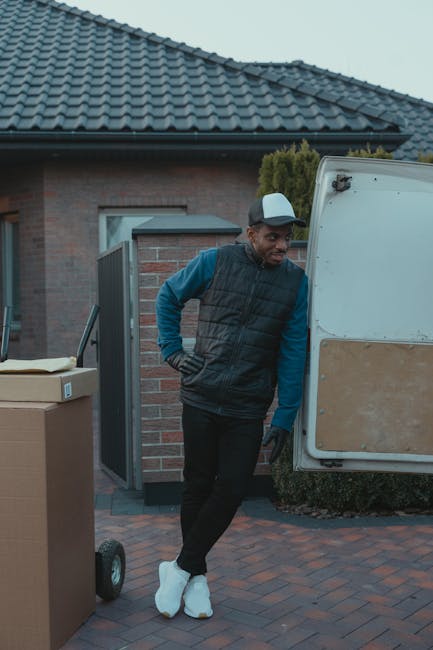 A man wearing a blue long-sleeve shirt, black padded vest, and white sneakers stands beside the open rear door of a white moving van parked on a brick driveway in front of a residential house with a tiled roof and brick walls. Several cardboard boxes and a wheeled trolley are visible nearby, indicating a home relocation process. The man is leaning slightly against the van, resting his head on his hand, as part of the loading or unloading of furniture and packing materials. In the background, green shrubbery and the house's exterior, including a window and a small gate, are visible. The scene is well-lit with natural daylight, and the image captures the logistical aspects of furniture transport and packing during a house move, as managed by Highbury Man and Van.