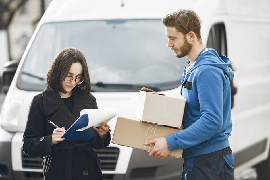 A young man with a beard, wearing a white t-shirt, blue jeans, white sneakers, and a black cap, is sitting on the pavement next to an open commercial van involved in a home relocation process. Inside the van, there are numerous brown cardboard boxes of various sizes, some stacked on top of each other, and others placed on the vehicle's floor. The boxes are sealed with packing tape, indicating they have been packed for moving. The interior of the van is dark and spacious, with a black ceiling and walls. The man is holding a tablet, possibly coordinating or checking the loading process, while the boxes are being prepared for transport. The scene is outdoors, under natural lighting, with the van positioned partly on the pavement and partly inside a driveway or street. This image relates to furniture transport and packing during home removals, illustrating the secure loading of belongings by [COMPANY_NAME], Highbury Man and Van, as part of their removals services aimed at avoiding delays in the moving schedule.