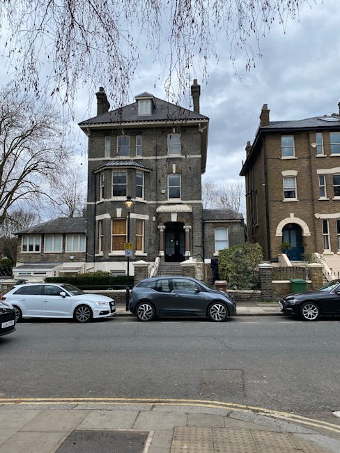 A three-storey Victorian-style house with a dark brick facade and a pitched roof, situated on a residential street. In front of the property, several parked cars, including a white sedan, a dark grey hatchback, and a black vehicle, line the pavement. The house features large bay windows on the ground and first floors, with the upper floor having smaller windows. There are stone steps leading to the front entrance, which is partially obscured by some shrubbery and a small gate. The surrounding environment includes leafless trees, indicating a winter or early spring season, and an overcast sky. Highbury Man and Van, a professional removals company, might use such properties during home relocation or furniture transport services, involving the packing, loading, and transportation of household belongings.