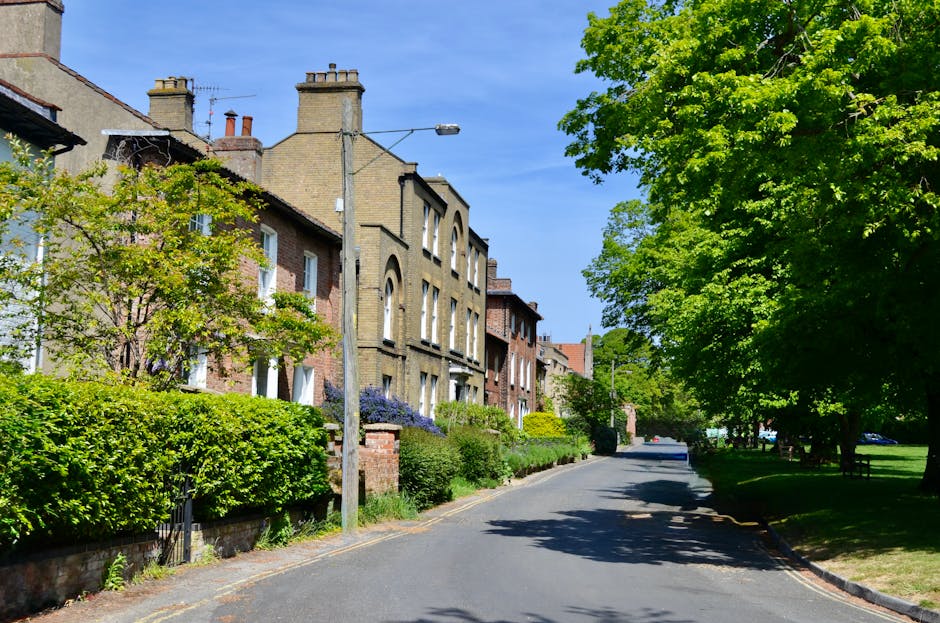 A three-storey Victorian-style house with a dark brick facade and a pitched roof, situated on a residential street. In front of the property, several parked cars, including a white sedan, a dark grey hatchback, and a black vehicle, line the pavement. The house features large bay windows on the ground and first floors, with the upper floor having smaller windows. There are stone steps leading to the front entrance, which is partially obscured by some shrubbery and a small gate. The surrounding environment includes leafless trees, indicating a winter or early spring season, and an overcast sky. Highbury Man and Van, a professional removals company, might use such properties during home relocation or furniture transport services, involving the packing, loading, and transportation of household belongings.