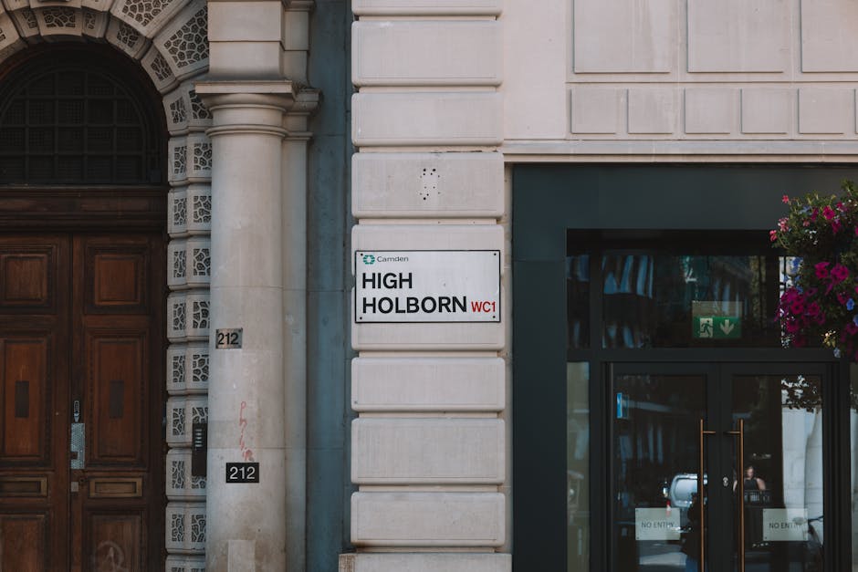 Image showing the exterior of a building with a white stone facade, featuring a large wooden double door to the left with decorative carvings and a silver handle. To the right of the door, there is a white rectangular sign with black and red text displaying the location as 'High Holborn' in the Camden area, with the postal code WC1. The sign is mounted on the building wall, which has a repeating pattern of square blocks. Adjacent to the sign, there is a glass door framed in dark metal, with a green emergency exit sign and a green arrow pointing downward visible through the glass. Potted flowers with vibrant pink and purple blooms are hanging near the top right corner of the glass entrance. The pavement area immediately outside the building appears accessible for loading, consistent with house or office relocations. Highbury Man and Van’s services related to removals and furniture transport are relevant for the context of moving procedures that comply with local regulations, such as those specified by Islington Council.