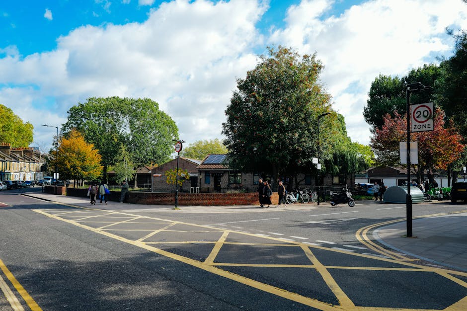 A wide street scene in a residential area during daytime, featuring a small brick building surrounded by mature trees with green and autumnal foliage. The sky is partly cloudy with patches of blue visible. Several pedestrians are walking along the sidewalk, some carrying bags, while a few bicycles and scooters are parked near the building. In the foreground, there are yellow grid markings on the road, indicating a restricted parking or loading zone, with a black van partially visible on the right side of the image. Street signage is visible, including a round 20mph speed limit sign and a zone sign. Highbury Man and Van's vehicle, likely a van used for home relocation and furniture transport services, is present nearby, possibly in the process of loading or unloading items as part of a house removal or moving logistics operation. The scene exemplifies typical urban moving activity, with objects such as boxes, furniture coverings, and packing materials potentially involved in the process, set within a well-maintained neighborhood environment.