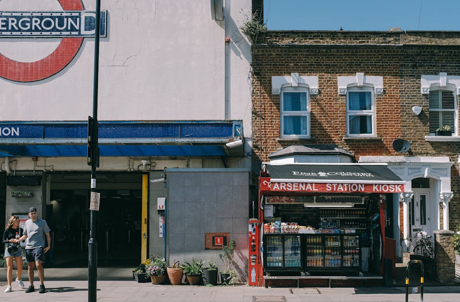 A street view showing a small kiosk with a red sign reading 'Arsenal Station Kiosk' situated on the pavement in front of residential buildings with brick facades and white window frames. To the left of the kiosk, there is a building with a white exterior and a blue signage indicating an underground station entrance. Two people, a man and a woman, are walking on the sidewalk near the entrance, carrying small bags. The scene is captured during daylight with clear skies, with shadows cast on the buildings and pavement. The image reflects a typical urban environment associated with house removals and furniture transport, where loading and packing activities could take place nearby during the house relocation process, and Highbury Man and Van may assist with such local moving logistics.