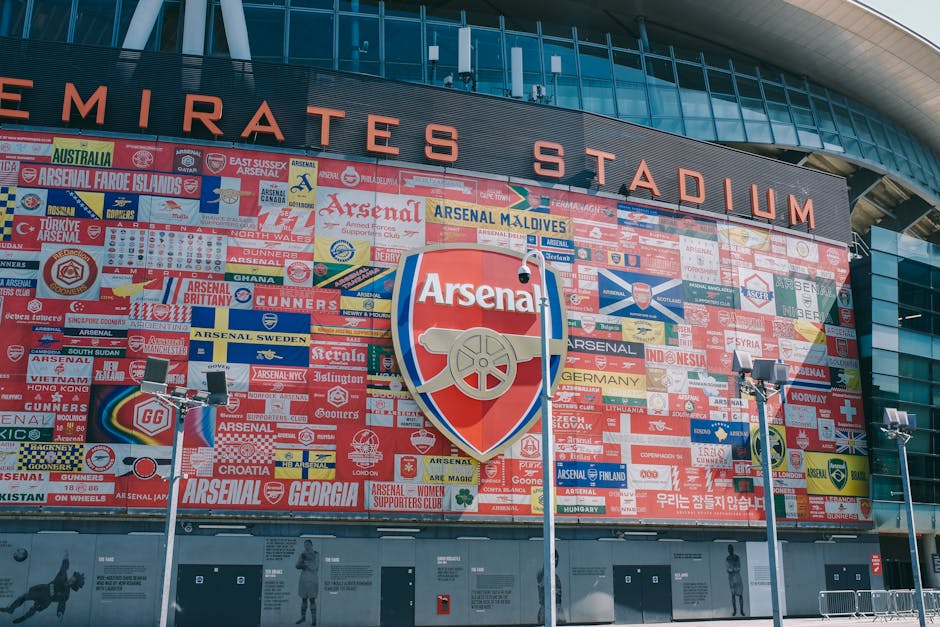 A large exterior view of Emirates Stadium, home to Arsenal Football Club, featuring a prominent wall covered with numerous colourful banners, stickers, and flags related to Arsenal. In the foreground, three tall floodlights on metal poles are visible, positioned on a paved area outside the stadium entrance. The stadium's modern glass and steel façade curves overhead, with the name 'EMIRATES STADIUM' displayed at the top. The image captures a daytime scene with clear lighting, providing a detailed view of the external decoration and signage associated with the football ground, relevant to house removals and moving services supporting events or relocations nearby. Highbury Man and Van might assist with home relocation or furniture transport in the area, ensuring smooth logistics for residential moves close to the stadium.