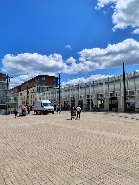 A street view showing a small kiosk with a red sign reading 'Arsenal Station Kiosk' situated on the pavement in front of residential buildings with brick facades and white window frames. To the left of the kiosk, there is a building with a white exterior and a blue signage indicating an underground station entrance. Two people, a man and a woman, are walking on the sidewalk near the entrance, carrying small bags. The scene is captured during daylight with clear skies, with shadows cast on the buildings and pavement. The image reflects a typical urban environment associated with house removals and furniture transport, where loading and packing activities could take place nearby during the house relocation process, and Highbury Man and Van may assist with such local moving logistics.